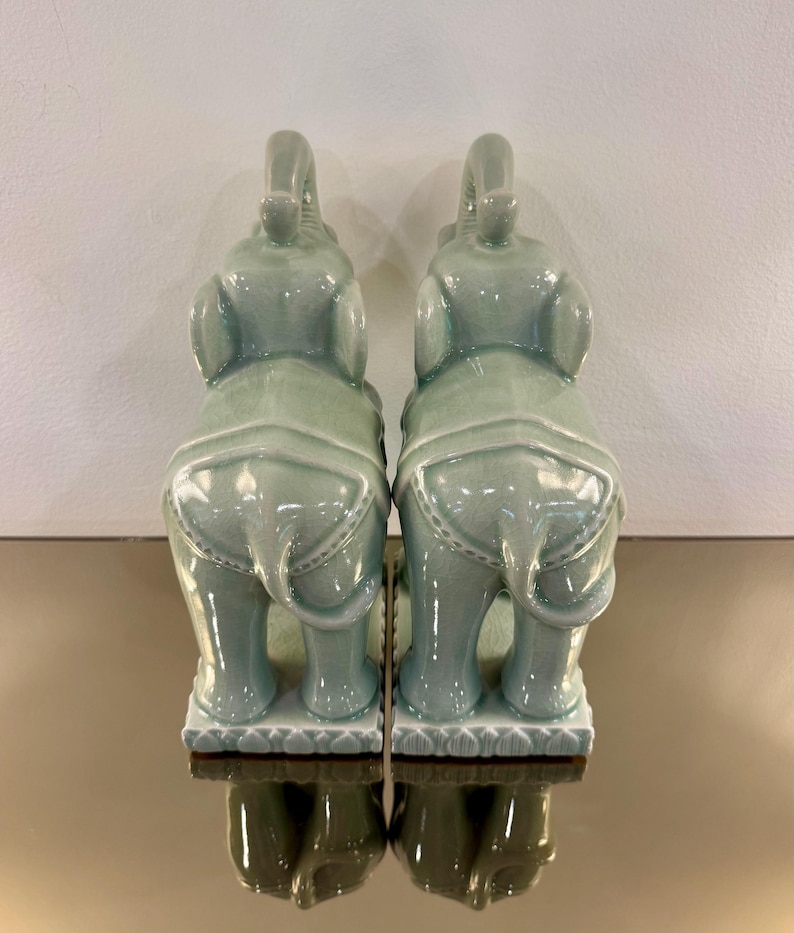 May include: A pair of celadon green ceramic elephant figurines standing on rectangular bases. Each elephant features a raised trunk, large ears, and a decorative saddle blanket pattern on its back. The glossy glaze has a subtle crackle finish.