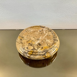 May include: A circular, polished stone tray featuring a mottled, earth-toned surface with beige, tan, brown, and grey fossilized inclusions. The tray has a smooth, rounded edge and a flat top surface, showcasing natural stone patterns throughout.