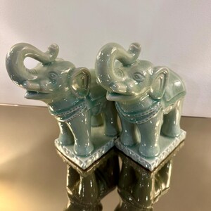May include: A pair of matching ceramic elephant figurines in a glossy celadon green glaze. Each elephant stands on a rectangular base with a decorative border, featuring an upturned trunk, detailed ears, and a saddle-like harness across the back.