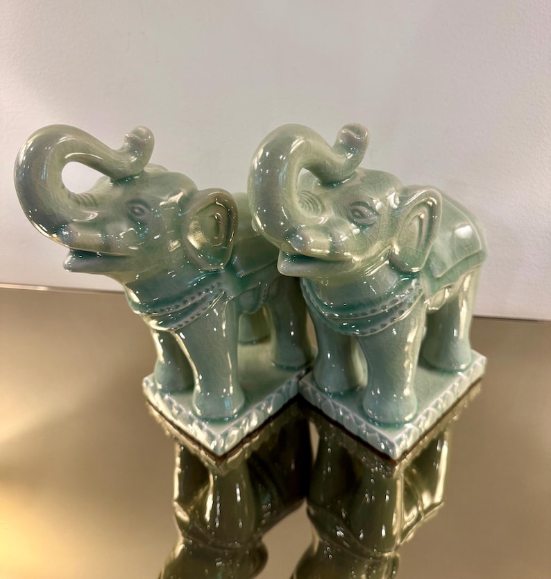 May include: A pair of matching ceramic elephant figurines in a glossy celadon green glaze. Each elephant stands on a rectangular base with a decorative border, featuring an upturned trunk, detailed ears, and a saddle-like harness across the back.