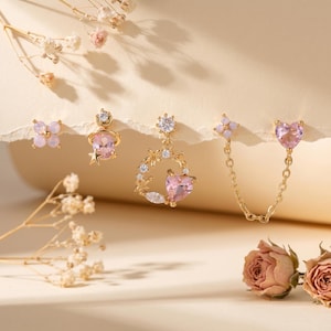 May include: An assortment of gold-toned earrings with pink and clear gemstones. The earrings include studs, dangles, and a chain-linked pair. Floral and heart-shaped designs are featured, with a soft, neutral background and dried flowers.