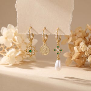 May include: Three gold-toned earrings with different designs. One features a starburst with a green center, another a coin, and the third a cross with a dangling pearl. All have small, clear stones around the hoops.
