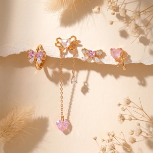 May include: A collection of gold-toned jewelry featuring bow and heart designs. The set includes a ring, earrings, and a pendant necklace. The jewelry incorporates pink and clear gemstones, creating a delicate and feminine aesthetic.