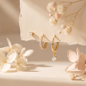May include: A set of three gold earrings with sparkling clear gemstones. The set includes a small stud, a hoop earring with gemstones, and a hoop earring with a dangling gemstone. The earrings are displayed on a cream-colored background with dried flowers.
