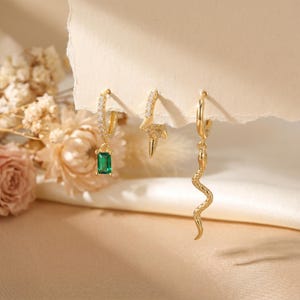Puede incluir: Tres pendientes dorados. Uno presenta una piedra verde talla esmeralda, otro un diseño de rayo y el tercero un colgante de serpiente. Los pendientes se exhiben sobre un fondo suave y neutro con detalles florales.