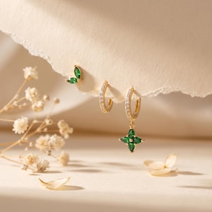 May include: A collection of gold-toned earrings. One pair features a four-leaf clover design with green gemstones. Another pair is a small hoop with clear stones. A third earring is a small leaf shape with green gemstones.