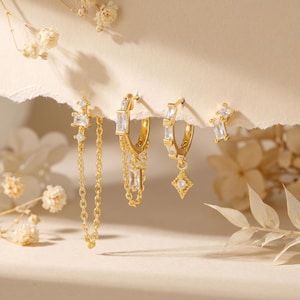 May include: Four pairs of gold-colored earrings with clear, rectangular and square-cut gemstones. One pair features a chain, another has a dangling gemstone, and a third pair is a simple stud. The earrings are displayed on a torn piece of paper.