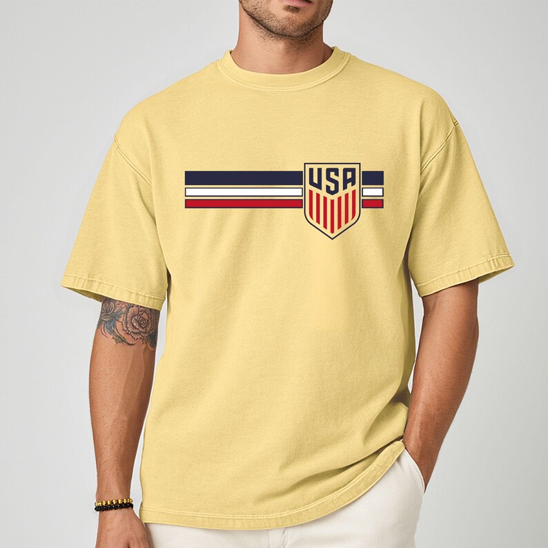Puede incluir: Una camiseta de manga corta de color amarillo p&aacute;lido con un gr&aacute;fico en el pecho. El dise&ntilde;o consta de tres franjas horizontales en azul marino, blanco y rojo, cruzadas por un emblema en forma de escudo con el texto USA en letras negritas.