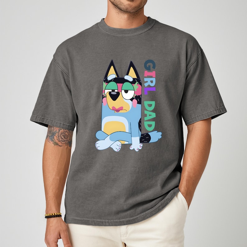 May include: Dark gray t-shirt featuring a cartoon dog character in blue, black, and yellow. The text "GIRL DAD" is printed vertically in colorful letters. The shirt has short sleeves and a relaxed fit.