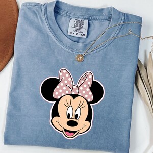 May include: A light blue Comfort Colors t-shirt with a Minnie Mouse graphic. The graphic shows Minnie's face with a pink polka dot bow. A gold necklace with a small pendant rests on the shirt. The shirt is size small.
