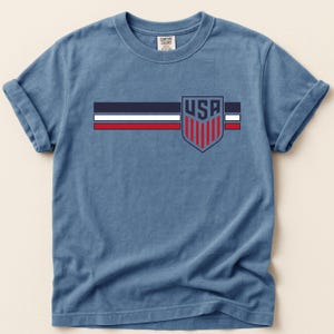 Puede incluir: Una camiseta azul de manga corta con un gr&aacute;fico en el pecho que presenta tres franjas horizontales en azul marino, blanco y rojo. A la derecha de las franjas hay un emblema de escudo con el texto USA sobre franjas rojas verticales en azul.