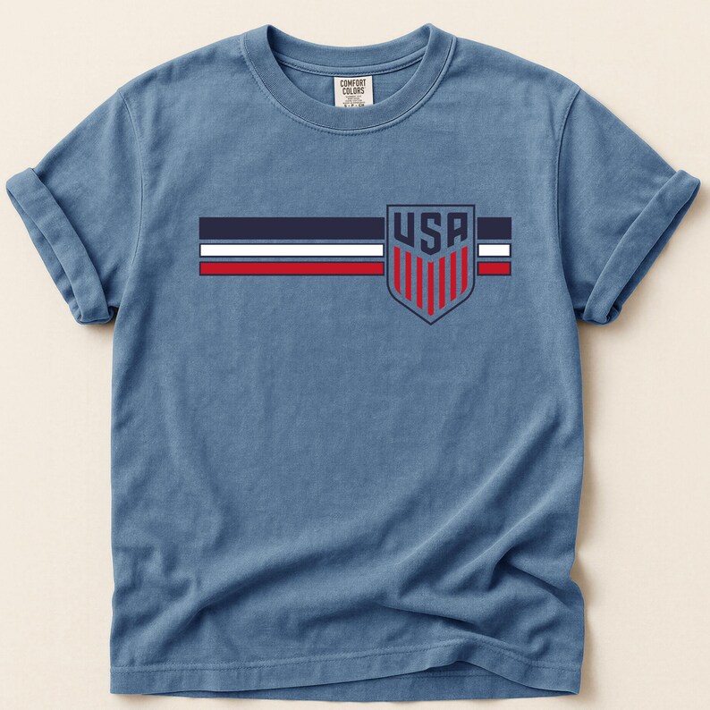 Puede incluir: Una camiseta azul de manga corta con un gr&aacute;fico en el pecho que presenta tres franjas horizontales en azul marino, blanco y rojo. A la derecha de las franjas hay un emblema de escudo con el texto USA sobre franjas rojas verticales en azul.