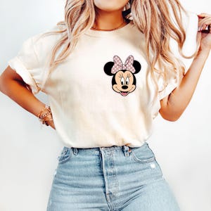 Op de afbeelding: Cr&egrave;mekleurig T-shirt met een Minnie Mouse-afbeelding. De afbeelding toont Minnie's gezicht met een roze strik met witte stippen. Het shirt heeft korte mouwen en een relaxte pasvorm, geschikt voor casual wear.