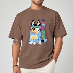May include: Brown t-shirt featuring a cartoon dog character in blue, black, and yellow, with the text "GIRL DAD" in colorful letters. The shirt has short sleeves and a relaxed fit.