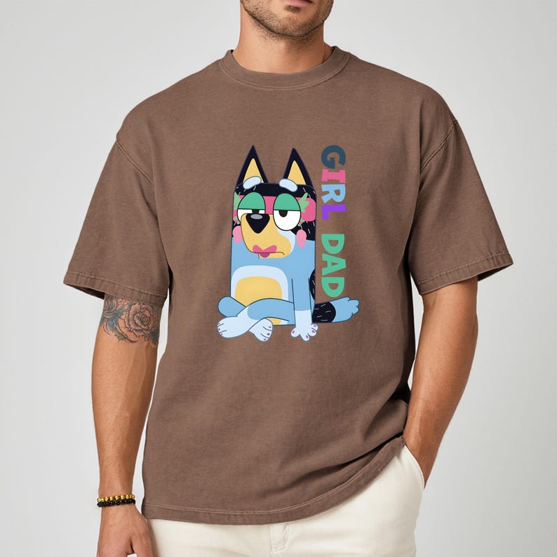 May include: Brown t-shirt featuring a cartoon dog character in blue, black, and yellow, with the text "GIRL DAD" in colorful letters. The shirt has short sleeves and a relaxed fit.