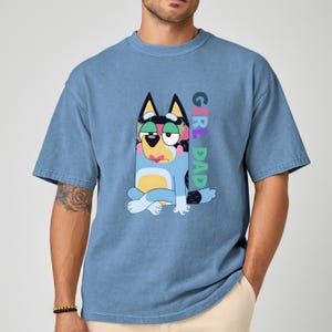 May include: Blue t-shirt featuring a cartoon dog character with the text "GIRL DAD" in colorful letters. The dog has blue fur, a yellow belly, and is sitting with its legs crossed. The shirt is a solid blue color.
