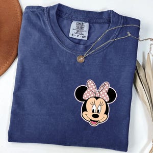 Op de afbeelding: Een marineblauw Comfort Colors T-shirt met een Minnie Mouse-afbeelding en een roze strik met stippen. Een gouden ketting met hanger ligt op het gevouwen T-shirt. Het label luidt S, P, CH.