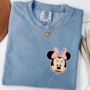 Op de afbeelding: Een blauw Comfort Colors T-shirt met een Minnie Mouse-afbeelding met een roze strik met stippen. Een gouden ketting met een hanger ligt op het shirt. Het label luidt "Comfort Colors" en "S - P - CH".