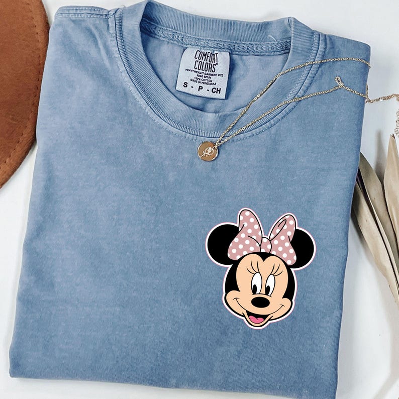 Op de afbeelding: Een blauw Comfort Colors T-shirt met een Minnie Mouse-afbeelding met een roze strik met stippen. Een gouden ketting met een hanger ligt op het shirt. Het label luidt "Comfort Colors" en "S - P - CH".