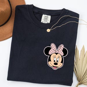 Op de afbeelding: Marineblauw T-shirt met een Minnie Mouse-afbeelding met een roze stippenstrik. Het shirt heeft een ronde hals en een klein label op de kraag. Een gouden ketting en een bruine hoed zijn ook zichtbaar.