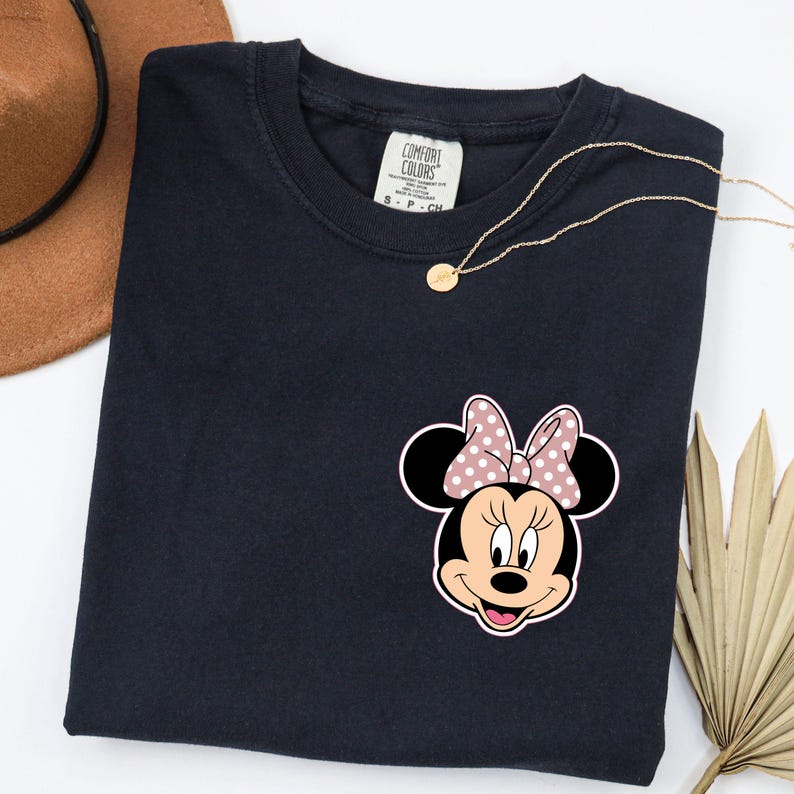 Op de afbeelding: Marineblauw T-shirt met een Minnie Mouse-afbeelding met een roze stippenstrik. Het shirt heeft een ronde hals en een klein label op de kraag. Een gouden ketting en een bruine hoed zijn ook zichtbaar.