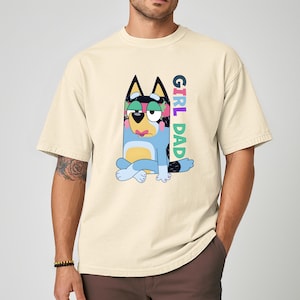 May include: Cream-colored t-shirt featuring a cartoon dog character with the text "GIRL DAD" in colorful letters. The dog is blue, with black ears and a sad expression. The shirt has short sleeves and a relaxed fit.