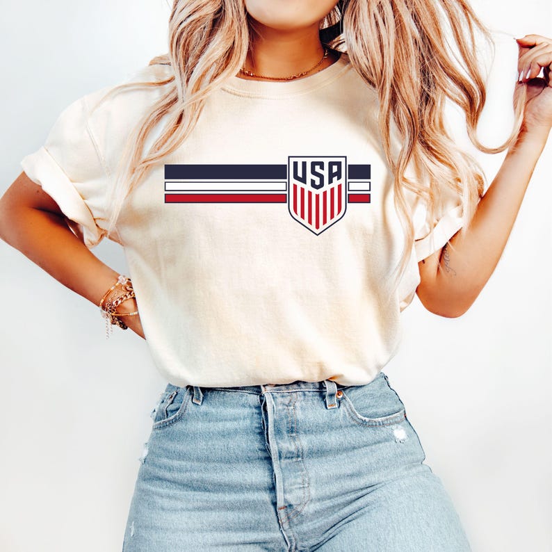 Puede incluir: Una camiseta de manga corta de color crema con un gr&aacute;fico en el pecho. El dise&ntilde;o consta de tres franjas horizontales en azul marino, blanco y rojo, cruzadas por un emblema en forma de escudo que contiene el texto USA en letras negritas.