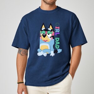 May include: Navy blue t-shirt featuring a cartoon dog character in blue, black, yellow, and pink. The dog is sitting with its legs crossed. The text "GIRL DAD" is printed vertically on the right side of the shirt.