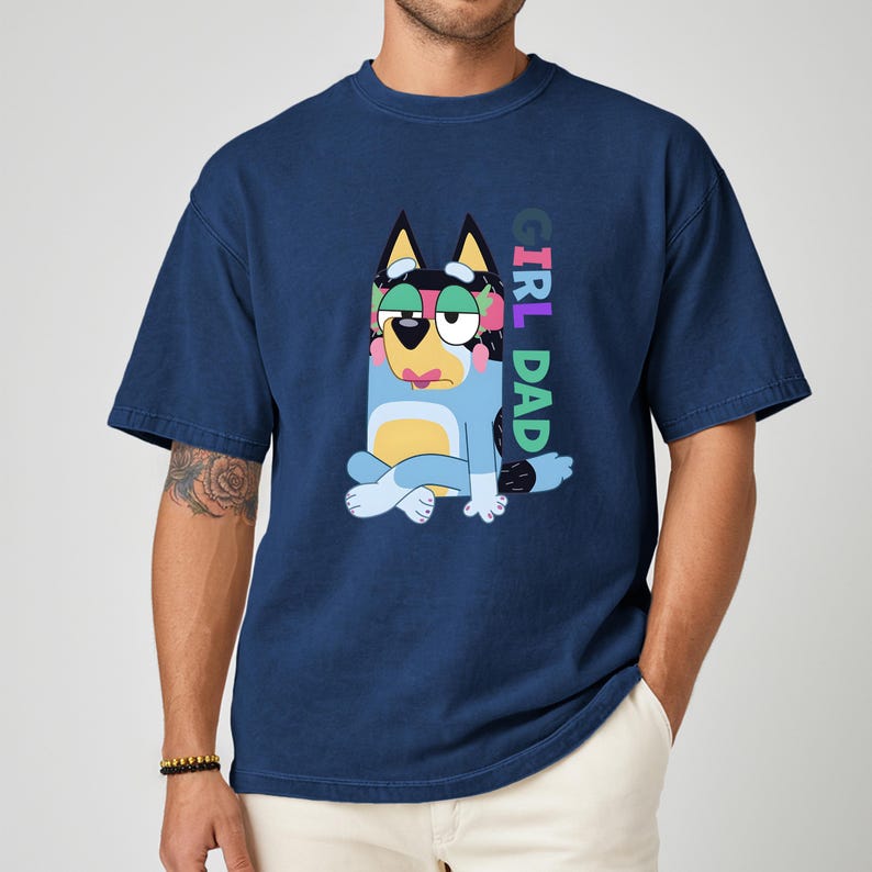 May include: Navy blue t-shirt featuring a cartoon dog character in blue, black, yellow, and pink. The dog is sitting with its legs crossed. The text "GIRL DAD" is printed vertically on the right side of the shirt.