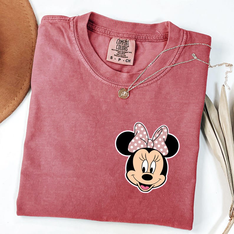 Op de afbeelding: Een stoffig roze Comfort Colors T-shirt met een Minnie Mouse-afbeelding en een roze stippenstrik. Een gouden ketting met een hanger ligt op het shirt. Het shirt heeft een ronde hals en korte mouwen. Een bruine hoed is gedeeltelijk zichtbaar.