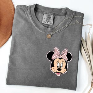 Op de afbeelding: Een grijze Comfort Colors t-shirt met een Minnie Mouse-afbeelding met een roze strik met stippen. Een zilveren ketting met een ronde hanger ligt over het gevouwen shirt op een witte ondergrond.