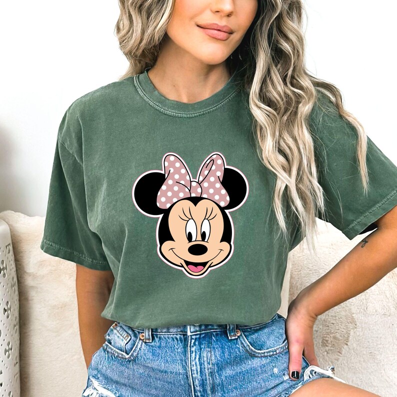 May include: Green t-shirt with a Minnie Mouse graphic. The graphic shows Minnie's face with a pink polka dot bow. The shirt has short sleeves and a crew neck. The shirt is made of a soft material.