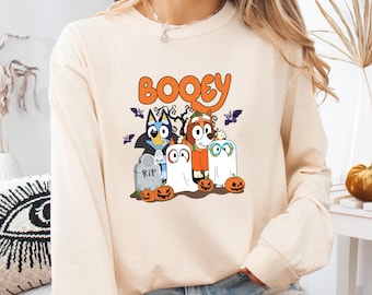 May include: A cream-colored long-sleeve sweatshirt featuring a graphic print with the text BOOEY in orange letters. The design includes two cartoon dogs dressed in Halloween costumes, ghosts, bats, a tombstone marked RIP, and several jack-o'-lanterns.