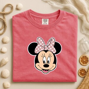 May include: A coral-coloured t-shirt featuring a Minnie Mouse graphic. The graphic shows Minnie's face with a polka-dot bow. The shirt has a crew neck and short sleeves. The tag reads "Comfort Colors".