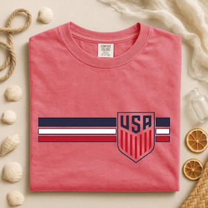 Puede incluir: Una camiseta de cuello redondo color coral con un dise&ntilde;o gr&aacute;fico horizontal de tres franjas en azul marino, blanco y rojo. A la derecha, un emblema en forma de escudo muestra el texto USA en azul marino sobre franjas verticales rojas.