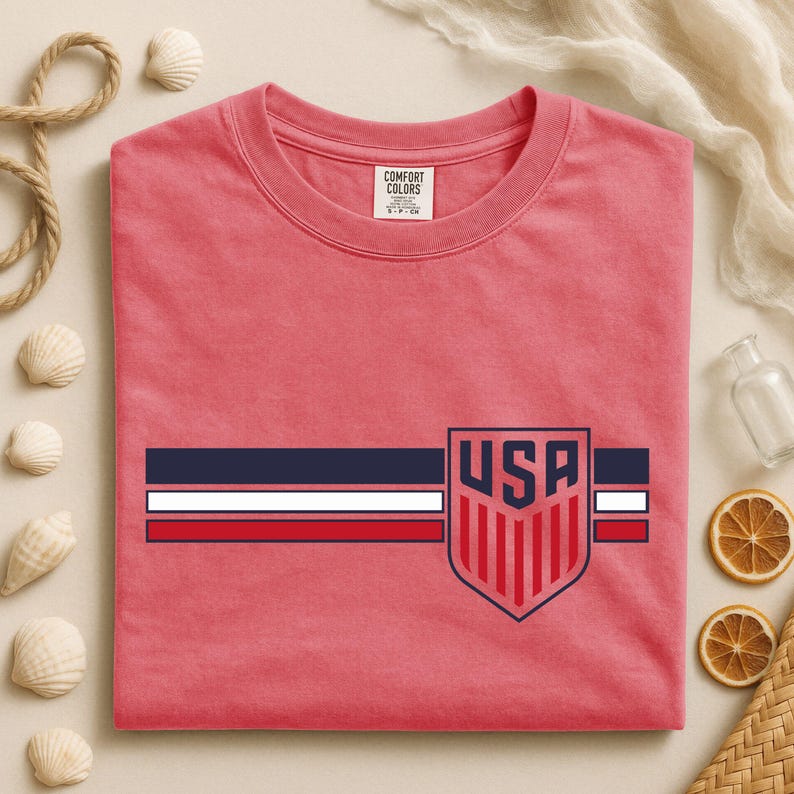Puede incluir: Una camiseta de cuello redondo color coral con un dise&ntilde;o gr&aacute;fico horizontal de tres franjas en azul marino, blanco y rojo. A la derecha, un emblema en forma de escudo muestra el texto USA en azul marino sobre franjas verticales rojas.