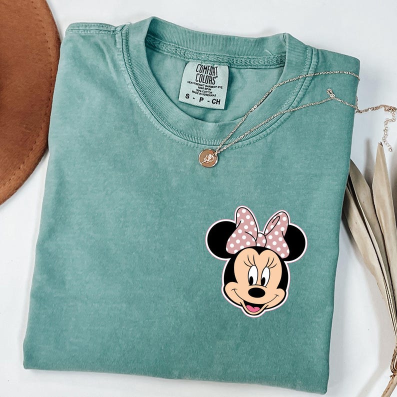 Op de afbeelding: Een zeegroen Comfort Colors T-shirt met een Minnie Mouse-afbeelding en een roze strik met stippen. Een zilveren ketting met hanger ligt op het gevouwen T-shirt.