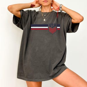 Puede incluir: Una camiseta de manga corta en color gris carb&oacute;n con un dise&ntilde;o gr&aacute;fico horizontal de rayas azul marino, blancas y rojas. En el lado derecho del pecho, un emblema en forma de escudo contiene el texto USA en letras azul marino en negrita.