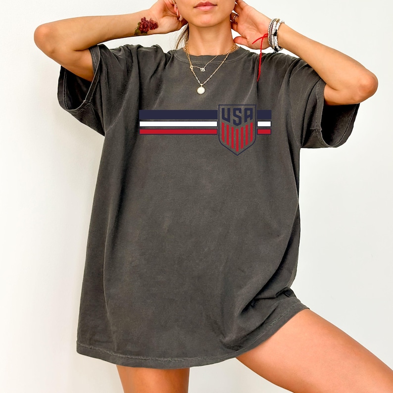 Puede incluir: Una camiseta de manga corta en color gris carb&oacute;n con un dise&ntilde;o gr&aacute;fico horizontal de rayas azul marino, blancas y rojas. En el lado derecho del pecho, un emblema en forma de escudo contiene el texto USA en letras azul marino en negrita.