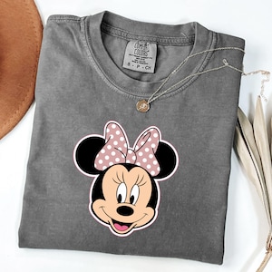 May include: A grey Comfort Colors t-shirt with a Minnie Mouse graphic. The graphic shows Minnie's face with a pink polka dot bow. A gold necklace with a pendant is on the shirt.