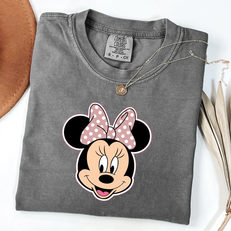 May include: A grey Comfort Colors t-shirt with a Minnie Mouse graphic. The graphic shows Minnie's face with a pink polka dot bow. A gold necklace with a pendant is on the shirt.