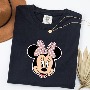 May include: A navy blue t-shirt with a Minnie Mouse graphic. The graphic shows Minnie's face with a pink polka dot bow. A gold necklace and a brown hat are also visible.
