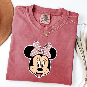 May include: A dusty rose Comfort Colors t-shirt with a Minnie Mouse graphic and a polka dot bow. A gold necklace with a pendant rests on the shirt. The shirt is folded, with a brown hat visible in the upper left corner.