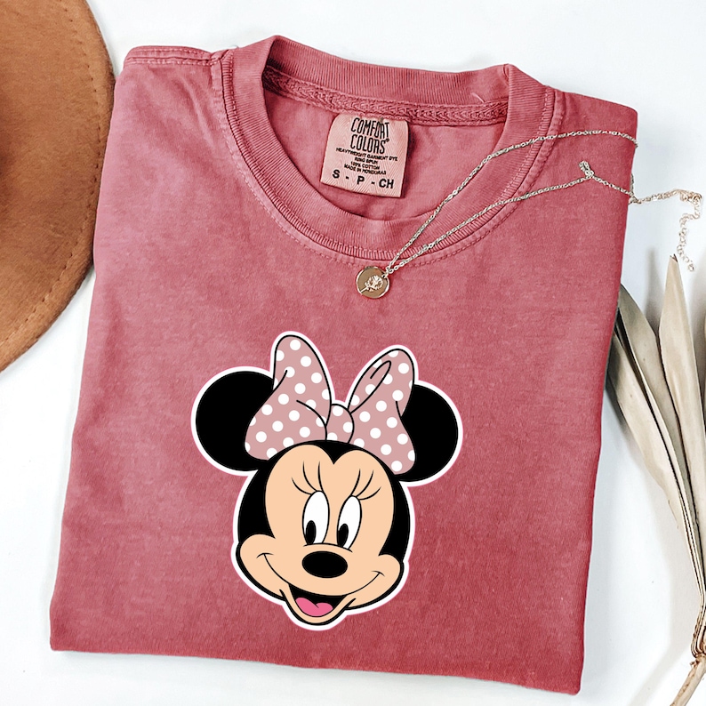 May include: A dusty rose Comfort Colors t-shirt with a Minnie Mouse graphic and a polka dot bow. A gold necklace with a pendant rests on the shirt. The shirt is folded, with a brown hat visible in the upper left corner.