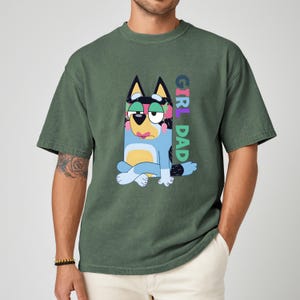 May include: Green t-shirt featuring a cartoon dog character with a colorful floral pattern and the text "GIRL DAD" in vertical rainbow letters. The dog is blue, yellow, and white, with a pink mouth and green eye makeup.