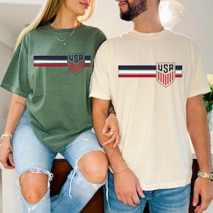 Puede incluir: Dos camisetas de manga corta y cuello redondo, una en verde oliva y otra en crema, cada una con un dise&ntilde;o gr&aacute;fico horizontal en el pecho. El gr&aacute;fico consta de tres rayas paralelas en azul marino, blanco y rojo, que terminan en un escudo con el texto USA.