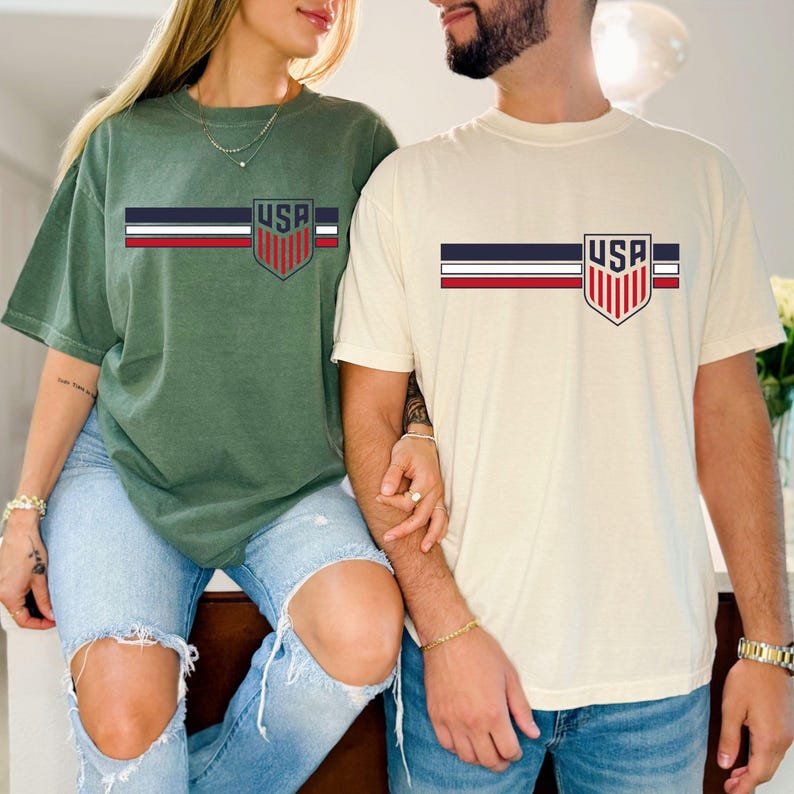 Puede incluir: Dos camisetas de manga corta y cuello redondo, una en verde oliva y otra en crema, cada una con un dise&ntilde;o gr&aacute;fico horizontal en el pecho. El gr&aacute;fico consta de tres rayas paralelas en azul marino, blanco y rojo, que terminan en un escudo con el texto USA.