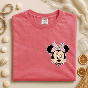 Op de afbeelding: Koraalrood T-shirt met een Minnie Mouse-afbeelding. De afbeelding toont Minnie's gezicht met een roze strik met stippen. Het shirt heeft een ronde hals en korte mouwen. Het label luidt "COMFORT COLORS".