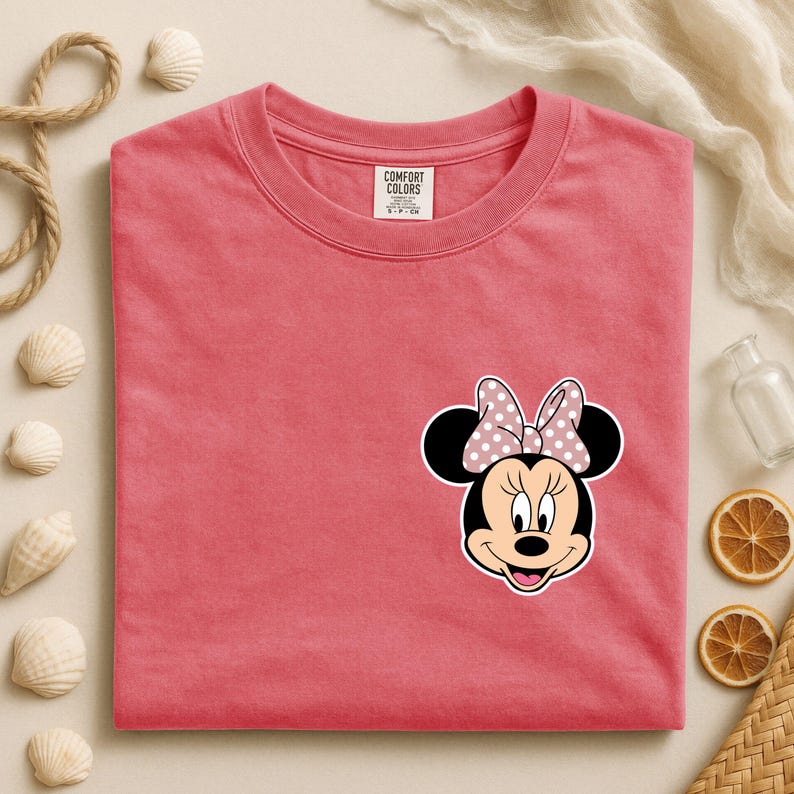 Op de afbeelding: Koraalrood T-shirt met een Minnie Mouse-afbeelding. De afbeelding toont Minnie's gezicht met een roze strik met stippen. Het shirt heeft een ronde hals en korte mouwen. Het label luidt "COMFORT COLORS".