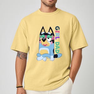 May include: Yellow t-shirt featuring a cartoon dog character with the text "GIRL DAD" in colorful letters. The dog is blue, black, and white, with a sad expression. The shirt is a solid yellow color.